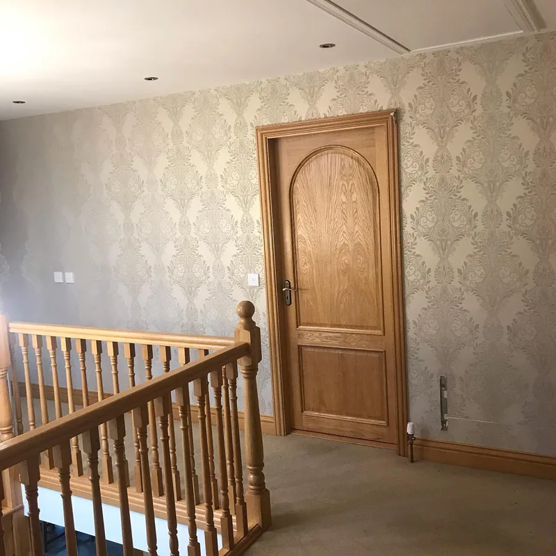 A landing hung with silver damask wallpaper beside an oak door and balustrade